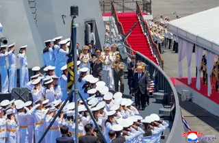 Kim Jong Un stands on the deck of a large grey warship, with naval equipment around him.