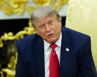 Donald Trump in a navy suit and red tie speaks while seated in the Oval Office