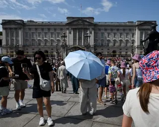 Crowds of tourists gather in front of Buckingham Palace on a sunny day, some using umbrellas for shade