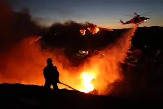 silhouette of firefighter standing behind roaring fire as helicopter overhead drops water