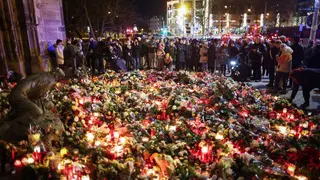 A large crowd gather at a red-and-white cordon at night, holding candles for a vigil. The front row mostly comprises women, all of whom wear large winter coats. It is night time. They are standing in front of a sea of flowers - many bouquets heaped on top of each other, stretching for several metres in every direction.