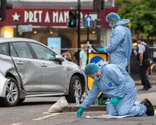 Two figures in blue protective clothing examine a road surface with a damaged silver car visible in the background.