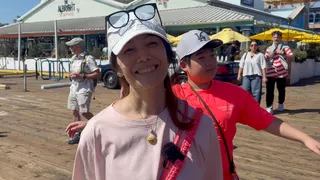 Maiko Asahi stands centre of framw wearing a baseball cap with eye glasses sitting on top of the brim. Her son stand behind her wearing a red shirt. They are on a boardwalk with a seafood restuarant in the background.