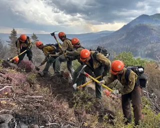 firefighters standing on hill holding axes as smoke rises in mountains in the background