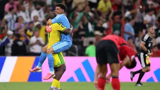 Ronwen Williams, wearing a blue goalkeeping kit, is held aloft in celebration after jumping into the arms of South Africa team-mate Mbekezeli Mbokazi, who is wearing a yellow kit with green trim. A despondent South Korea player in red shirt, black shorts and red socks can be seen out of focus in the foreground, while in the distance a black-clad referee's assistant, pink advertising hoardings and a crowd are visible