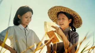 畫面中兩名女性站在田間，其中一人正向另一人念出一封信的內容。