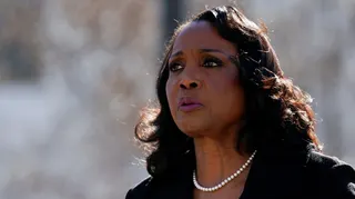 Lisa good is turned partially with a blurred grey background. She is wearing pearls, a black suit and appears to be speaking outside