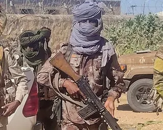 Armed fighters in camouflage and face coverings hold rifles in an outdoor area with vehicles and vegetation