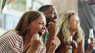A woman with long blonde hair cheers with two friends in what appears to be a sitting room. Her friends are holding bottles of beer. She is wearing a brown and white striped top.