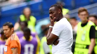 Pape Thiaw, visible from waist up wearing a white t-shirt, holds his left hand up to his chin in a thoughtful pose on the touchline during a football match. Out of focus in the background an assistant referee can be seen in an orange shirt, substitutes wear purple bibs and stewarding staff are in luminous yellow bibs