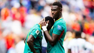 Lamine Camara of Senegal leans his face against the chest of team-mate Moussa Niakhate as he is comforted following the World Cup 2026 last-32 loss to Belgium in Seattle. Niakhate has his hands clasped around the back of Camara's head to pull him in. Both are visible from waist up and wearing their green Senegal jerseys with yellow and red trim. The background is a highly-blurred scene which includes one Belgium player seen from behind and the crowd