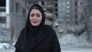 A woman looks directly at the camera. She is wearing black clothes including a headscarf.