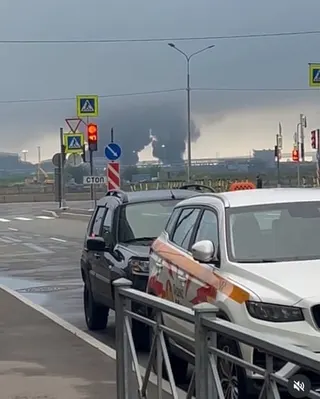Two plumes of black smoke visible on the horizon behind cars parked in a Russian industrial port zone.