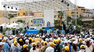 The Pope stands on a stage under a white tent. He is flanked by officials on the stage and in front of him are dozens of people wearing yellow and white hats.