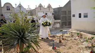 The Pope holds is holding flowers at a cemetery. Behind him are officials dressed in black suits and a row of white buildings with crosses on them.