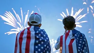 Two boys watch a fireworks display during a celebration of the US Army's 250th anniversary on Flag Day, June 14, 2025 in Washington DC. They are draped in USA flag and the sky in front of them is alight with fireworks