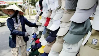 Caps bearing the logos of Polo, GAP, Prince and others hang in the foreground next to a woman wearing a blue blazer and a straw nón lá hat