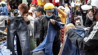 Women, including one wearing a bright yellow motorcycle helmet, look at pairs of jeans at an outdoor market