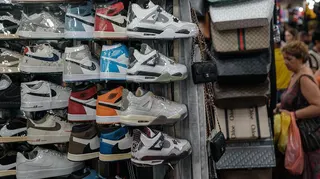 Plastic-wrapped Nike sneakers racked at a marketplace alongside handbags while people shop in the background