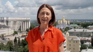 Sarah Rainsford wears a short-sleeved bright orange shirt. She reports in front of several buildings in Crimea