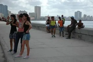 在古巴哈瓦那，阴天笼罩着大地，人们沿着马雷贡海滨长廊行走或驻足，背景是城市的天际线和海湾。