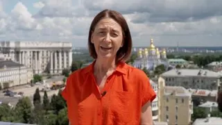Sarah Rainsford wears a short-sleeved bright orange shirt. She reports in front of several buildings in Crimea