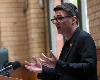 Andy Burnham stands before a lectern and microphones, speaking and gesturing emphatically with his hands