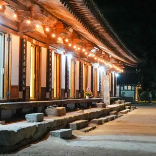 Traditional Korean temple building illuminated by warm lights at night, with a curved tiled roof, wooden columns, and stone steps lining a wide courtyard. Several visitors walk along the path beside the temple.