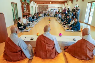 A large group of participants sit on floor cushions arranged in a wide semi-circle inside a temple hall with three monks at the front.
