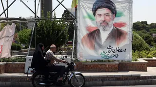 A man and a woman on a motorcycle pass a large portrait of Supreme Leader Ayatollah Mojtaba Khamenei on a street in Tehran.