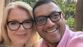 A smiling blonde woman wearing glasses is seen cuddled up next to a smiling man with short brown hair also wearing glasses in a selfie