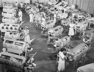 black and white photo of dozens of children inside metal cylindrical devices as nurses stand by observing