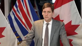 David Eby stand at a podium (unseen) with Canada's and British Columbia's flags behind him.