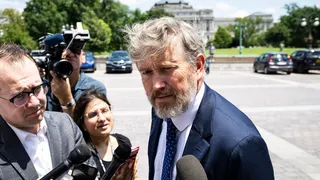 Man in a navy suit and blue tie speaks to reporters with multiple microphones; a cameraman films him as a domed government building and green trees are in the background.