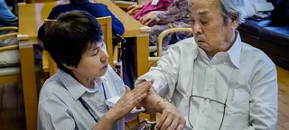 一名女护理员正在检查一位亚裔老年男子的手臂，地点似乎是在一家护理机构。