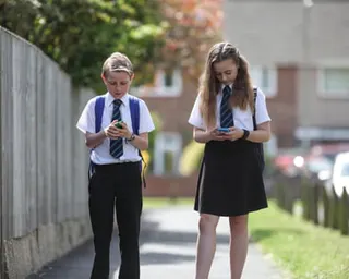 A girl and a boy in school uniform looking at their phones while walking.