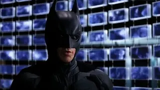 Batman stands in front of computer screens.