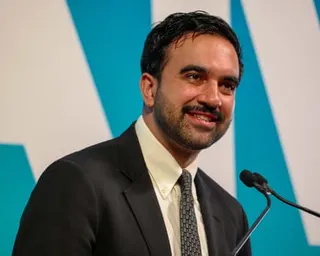 man in a dark suit and patterned tie smiles while speaking at a microphone
