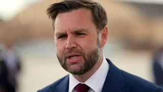 A close-up image of JD Vance talking to reporters. He wears a dark blue jacket, white shirt and dark red tie. He has short brown hair.