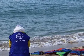 一名身穿蓝色 IOM 背心的人站在岸边，眺望着大海，沙滩上放着救生衣。