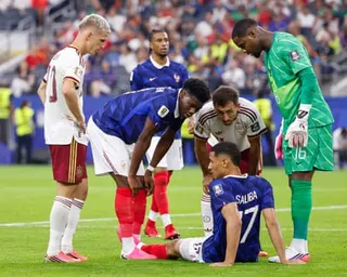 William Saliba sits on the pitch holding his leg while teammates and opponents look on with concern