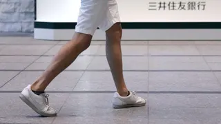 A pair of legs in white shorts and white sneakers walking inside an office building, in front of a low white sign that says "Sumitomo Mitsui Banking Corporation" in Japanese characters