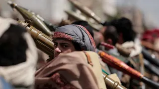 A man with a serious expression, wearing a head covering, holds what appears to be a bazooka over his shoulder, surrounded by other men holding the same weapons