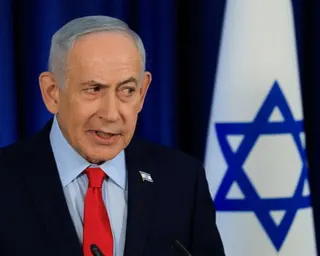 man in suit speaks into microphones, an israeli flag displayed behind him