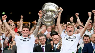 Two young men are standing with their backs to a large crowd in a stadium. They are both wearing white football tops and are holding the large, silver Sam Maguire trophy up between them. They are shouting and celebrating.