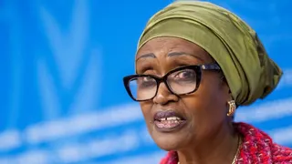 Winnie Byanyima wearing a khaki headwrap with a mic in front of her. the background is light blue