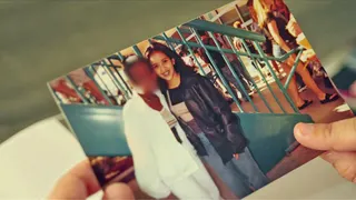A printed photograph being held in two hands. The photo shows Isabel as a teenager, smiling and posing with a friend whose face has been blurred. They are near dark blue metal railings in a busy public place. Isabel is wearing a dark jacket and jeans, while the blurred person is dressed in white.