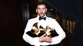Bad Bunny wears a white shirt with a black bow tie. He is holding numerous Grammy Awards.