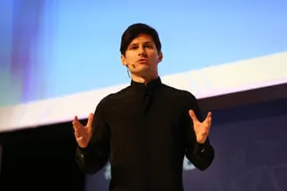 Pavel Durov gives a speech: he wears a headset microphone and holds his hands out in a gesticulation. He has short dark hair and is clean-shaven, and wears a black polo-neck jumper.