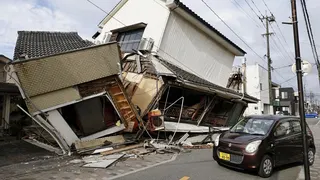 A small brown car sits on the street beside a collapsed house